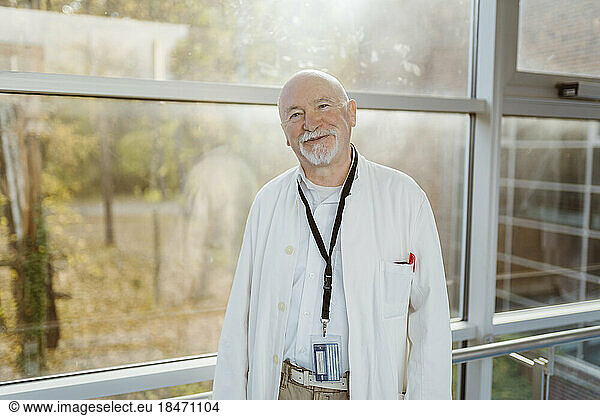 Portrait of smiling senior male doctor leaning on railing against window at hospital corridor