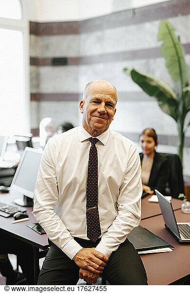 Portrait of smiling senior male lawyer sitting on desk in office