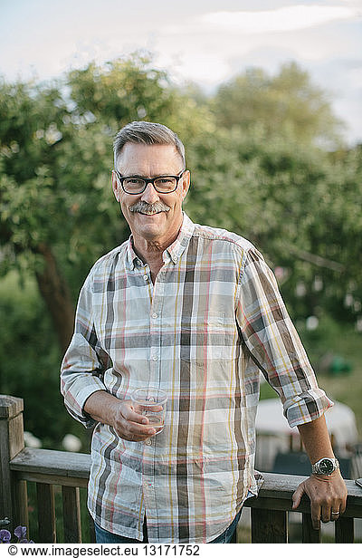 Portrait of smiling senior man holding drinking glass standing on porch