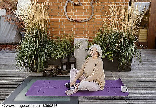 Portrait of smiling senior woman sitting on exercise mat at terrace