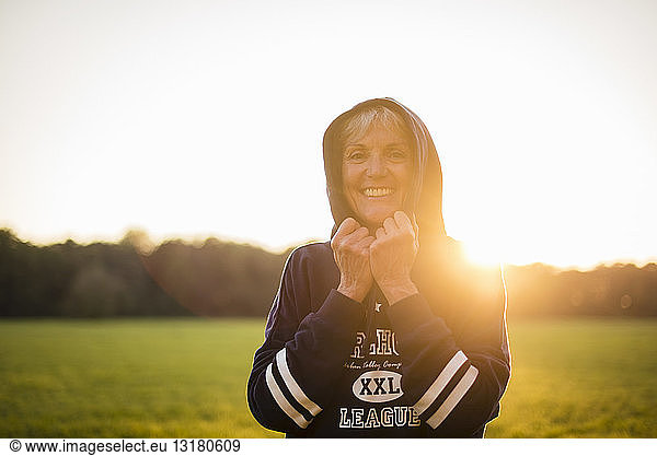 Portrait of smiling senior woman wearing a hoodie standing on rural meadow at sunset