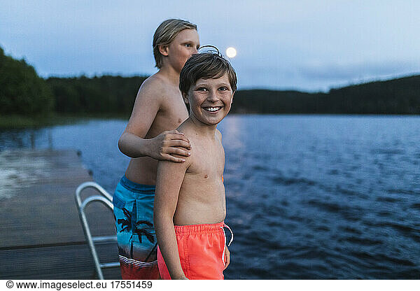Portrait of smiling shirtless boy standing with brother by sea at dusk