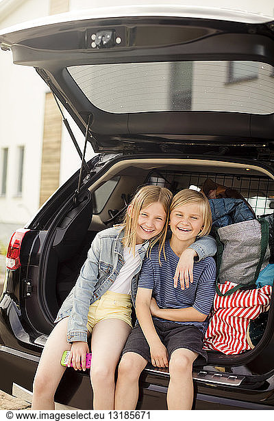 Portrait of smiling siblings sitting in car trunk against house