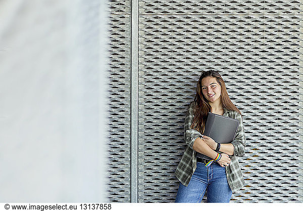 Portrait of smiling student holding file while standing against patterned wall in city