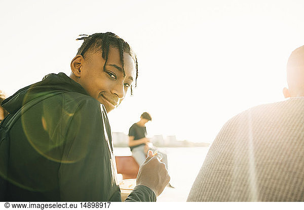 Portrait of smiling teenage boy holding meal while sitting by friend on promenade during sunny day