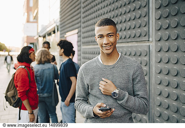Portrait of smiling teenage boy holding mobile phone while friends standing in background on street