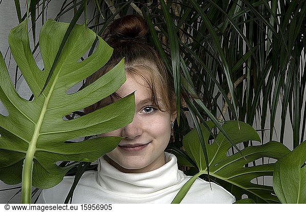 Portrait of smiling teenage girl hiding behind Monstera leaf