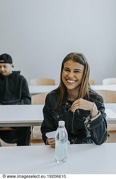 Portrait of smiling teenage girl sitting with smart phone and water bottle at desk in classroom