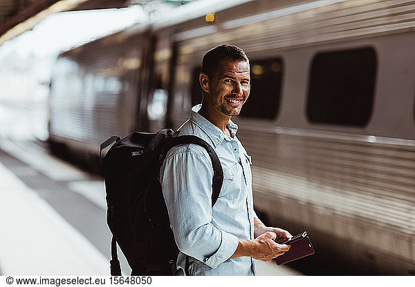 Portrait of smiling tourist with backpack holding smart phone on platform at train station