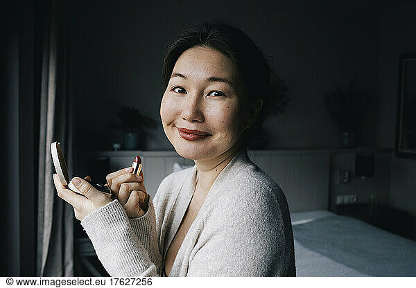 Portrait of smiling woman applying lipstick in bedroom at home