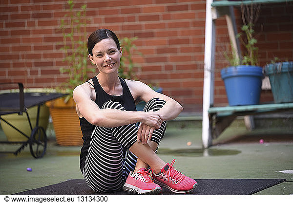 Portrait of smiling woman sitting on exercise mat against brick wall