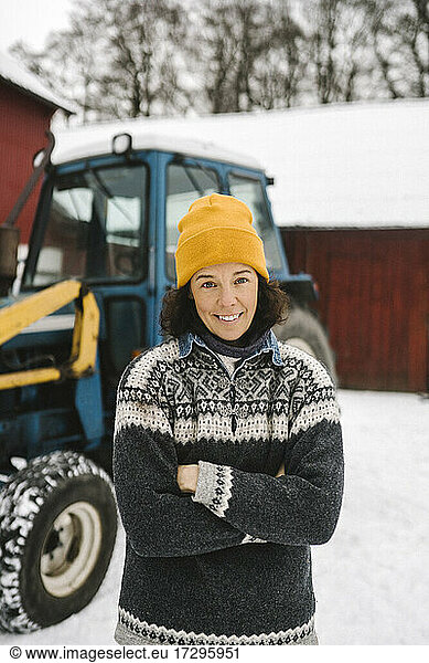 Portrait of smiling woman standing with arms crossed against tractor during winter