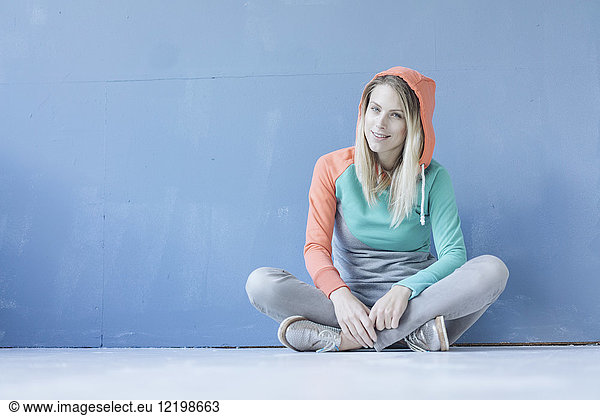 Portrait of smiling woman wearing hooded jacket sitting on the floor in front of blue wall