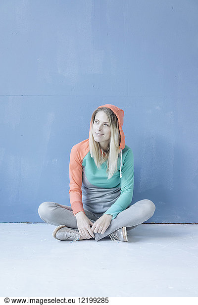 Portrait of smiling woman wearing hooded jacket sitting on the floor in front of blue wall