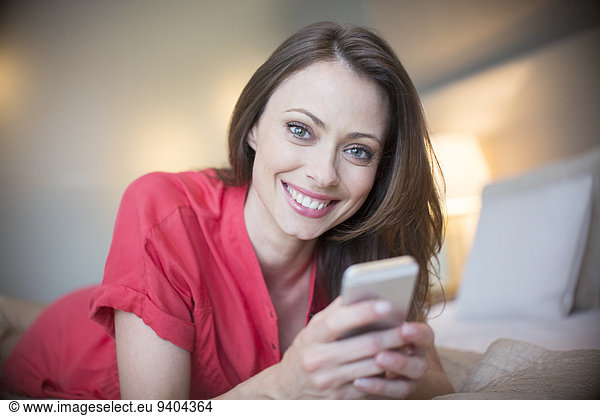 Portrait of smiling woman wearing red dress lying on bed with smartphone
