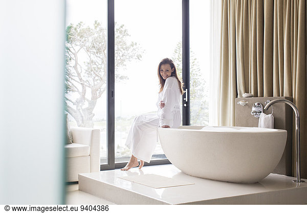 Portrait of smiling woman wearing white bathrobe  sitting on edge of bathtub in bathroom