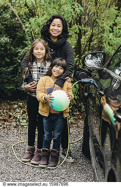 Portrait of smiling woman with daughters by electric car at charging station