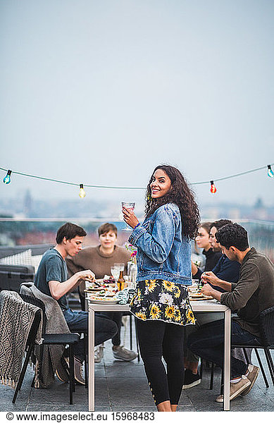 Portrait of smiling woman with drink standing by table during social gathering on terrace building