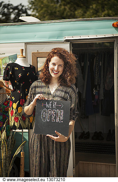 Portrait of smiling woman with opening board standing against camper van