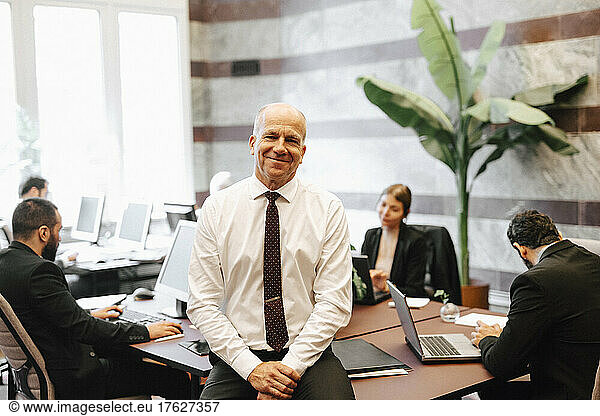 Portrait of smiling working senior male lawyer sitting on desk in office