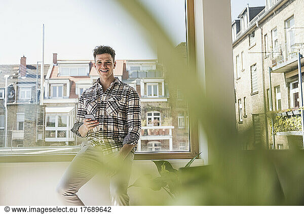 Portrait of smiling young businessman with smartphone at the window in office