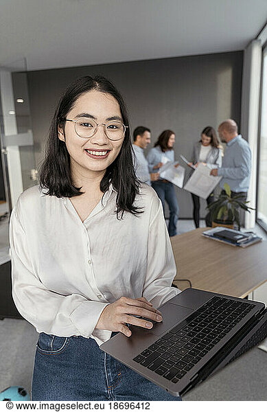 Portrait of smiling young businesswoman using laptop with colleagues in background in office