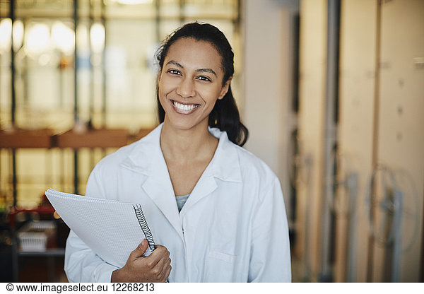 Portrait of smiling young chemistry student wearing lab coat standing with book in university