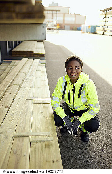 Portrait of smiling young female worker crouching by planks at lumber industry