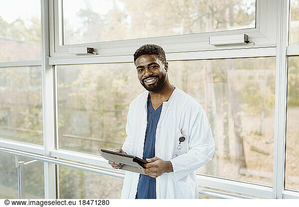 Portrait of smiling young male doctor holding digital tablet standing against window at hospital corridor