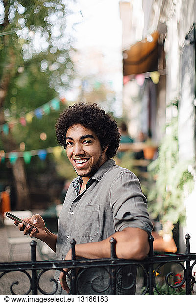 Portrait of smiling young man holding mobile phone while leaning on railing in backyard