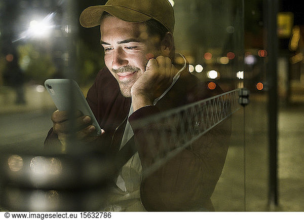 Portrait of smiling young man sitting at bus stop by night looking at cell phone  Lisbon  Portugal