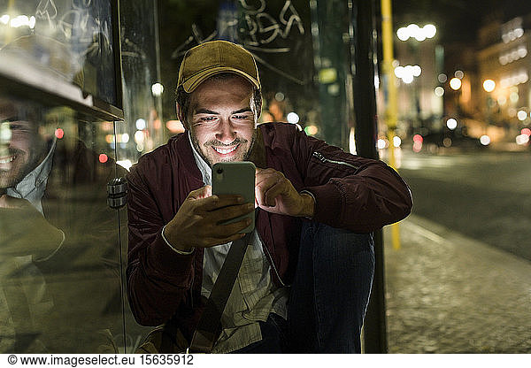 Portrait of smiling young man sitting at bus stop by night using cell phone  Lisbon  Portugal