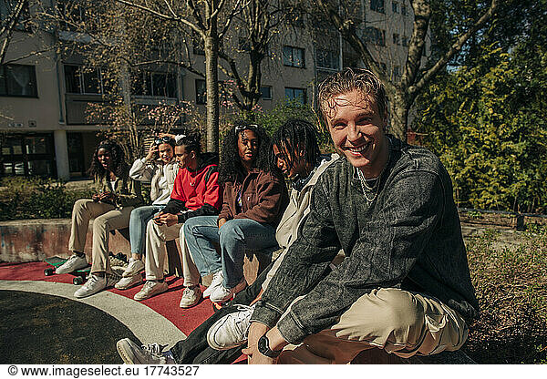 Portrait of smiling young man sitting with multiracial friends on retaining wall in playground