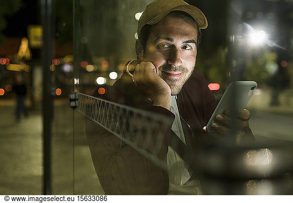Portrait of smiling young man with smartphone waiting at bus stop by night  Lisbon  Portugal