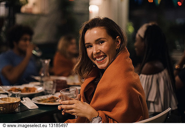 Portrait of smiling young woman holding drink while sitting at table during dinner party in backyard