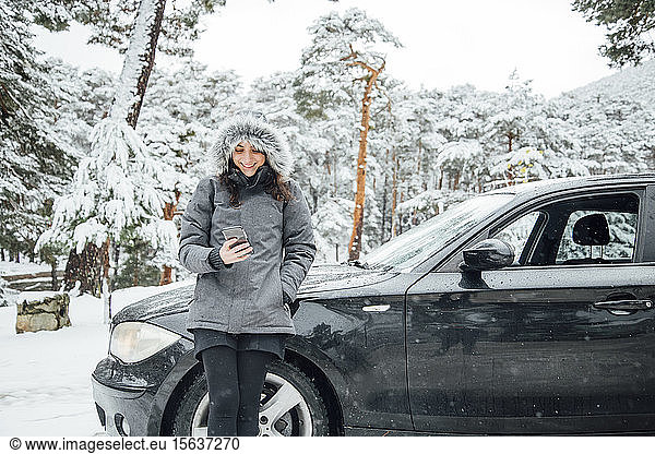 Portrait of smiling young woman leaning at parked car in winter forest looking at cell phone