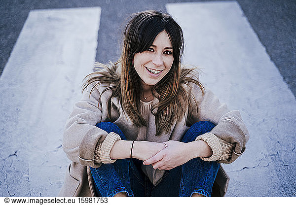 Portrait of smiling young woman sitting on zebra crossing