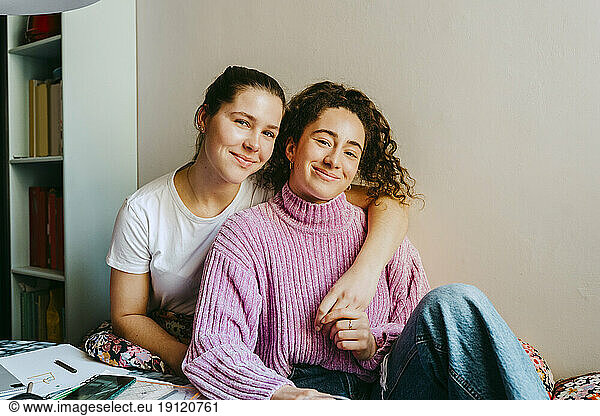 Portrait of smiling young woman with arm around friend sitting at home
