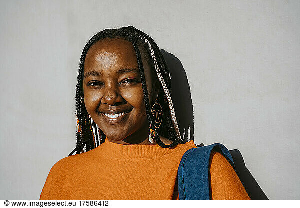 Portrait of smiling young woman with braided hairstyle against gray wall on sunny day
