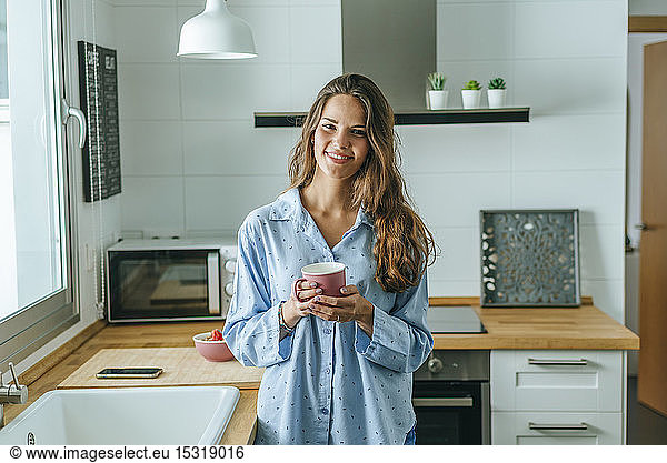 Portrait of smiling young woman with cup of coffee wearing pyjama in kitchen at home