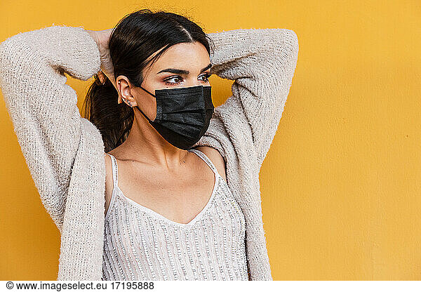 Portrait of Spanish brunette girl with face mask holding her hair on yellow background wall.