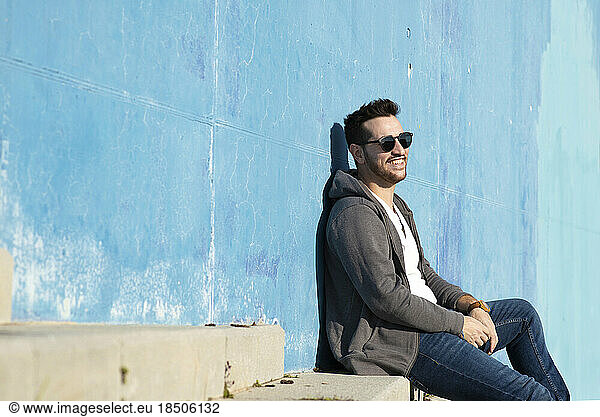 Portrait of stylish man sitting on stairs leaning on a blue wall while looking away and smiling