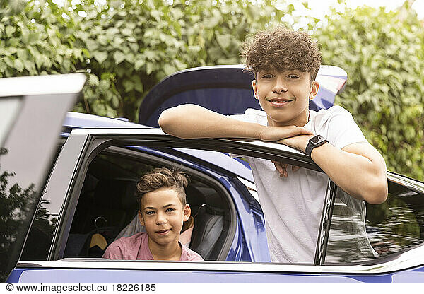 Portrait of teenage boy leaning on door with brother sitting in car