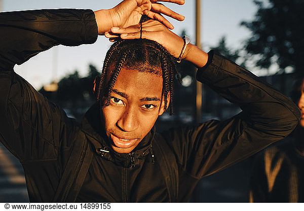 Portrait of teenage boy tying braided hair in city during sunset