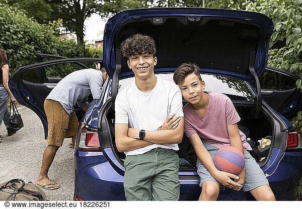 Portrait of teenage boy with arms crossed sitting by brother in car trunk
