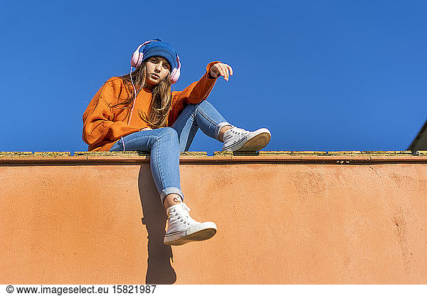 Portrait of teenage girl sitting on wall against sky listening music with headphones