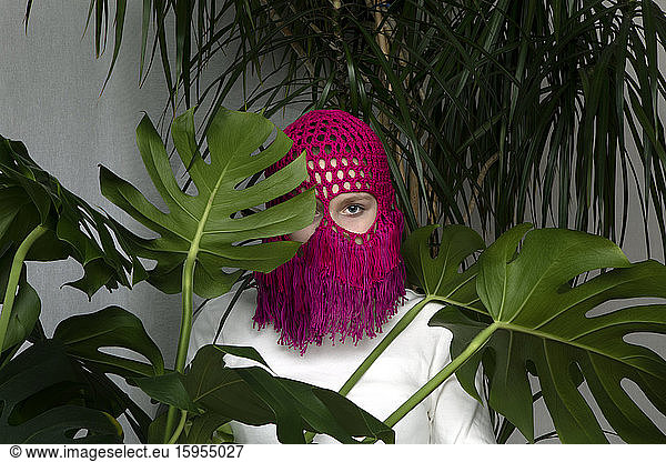 Portrait of teenage girl wearing crocheted pink headdress hiding behind Monstera leaves