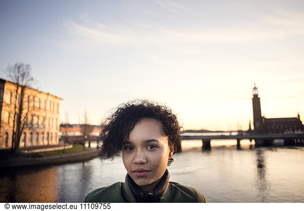 Portrait of teenage girl with curly short hair standing against canal in city
