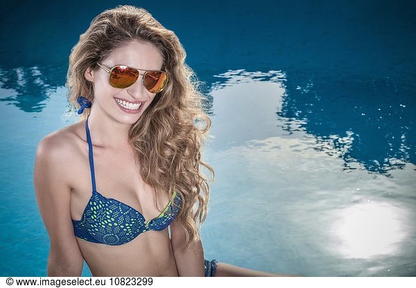 Portrait of teenage girl with long wavy hair sitting at poolside wearing bikini