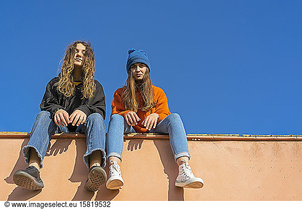 Portrait of teenage girls sitting on a wall against blue sky